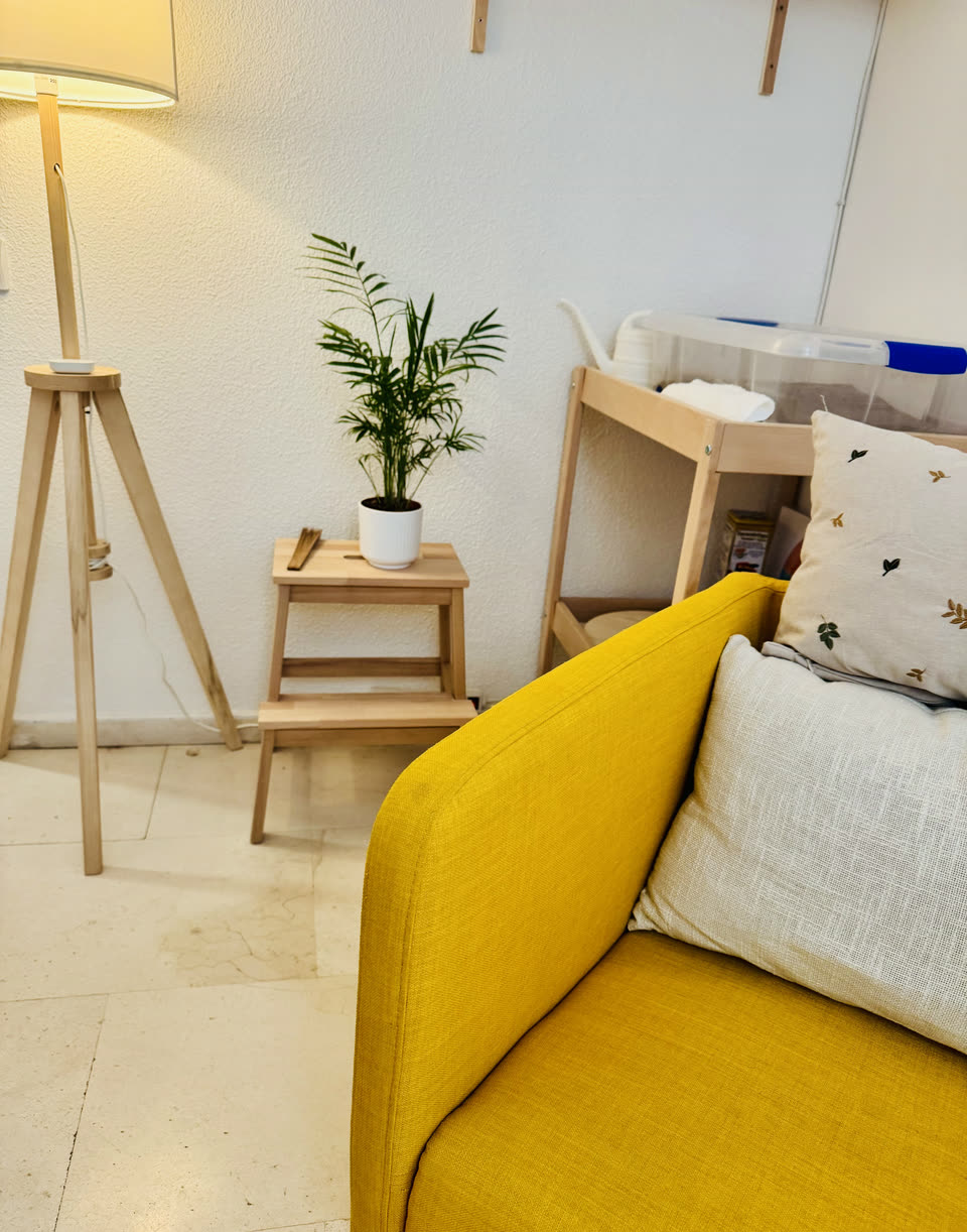 Rincón de la sala de consulta: sofá mostaza, lámpara de madera y planta junto a la pared.