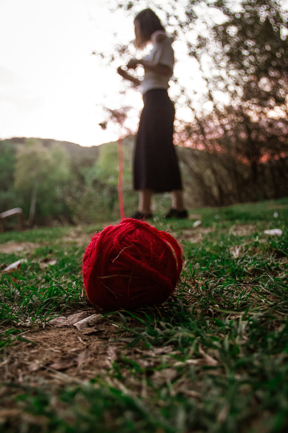 Una bola de lana roja con el hilo extendido en primer plano y una mujer de pie al fondo, en un campo al atardecer.